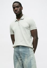 Svijetlosiva polo majica s ovratnikom na patentni zatvarač, s malim kvadratnim logotipom, uparena s izblijedjelim plavim trapericama s uzorkom. Glatka tekstura, ležerni dizajn.