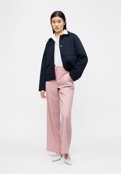 Γυναίκα στέκεται, φοράει μπλε σκούρο σακάκι, λευκό πουκάμισο, ροζ παντελόνι με φαρδιά μπατζάκια και λευκές μυτερές γόβες, μπροστά σε απλό ανοιχτό φόντο.