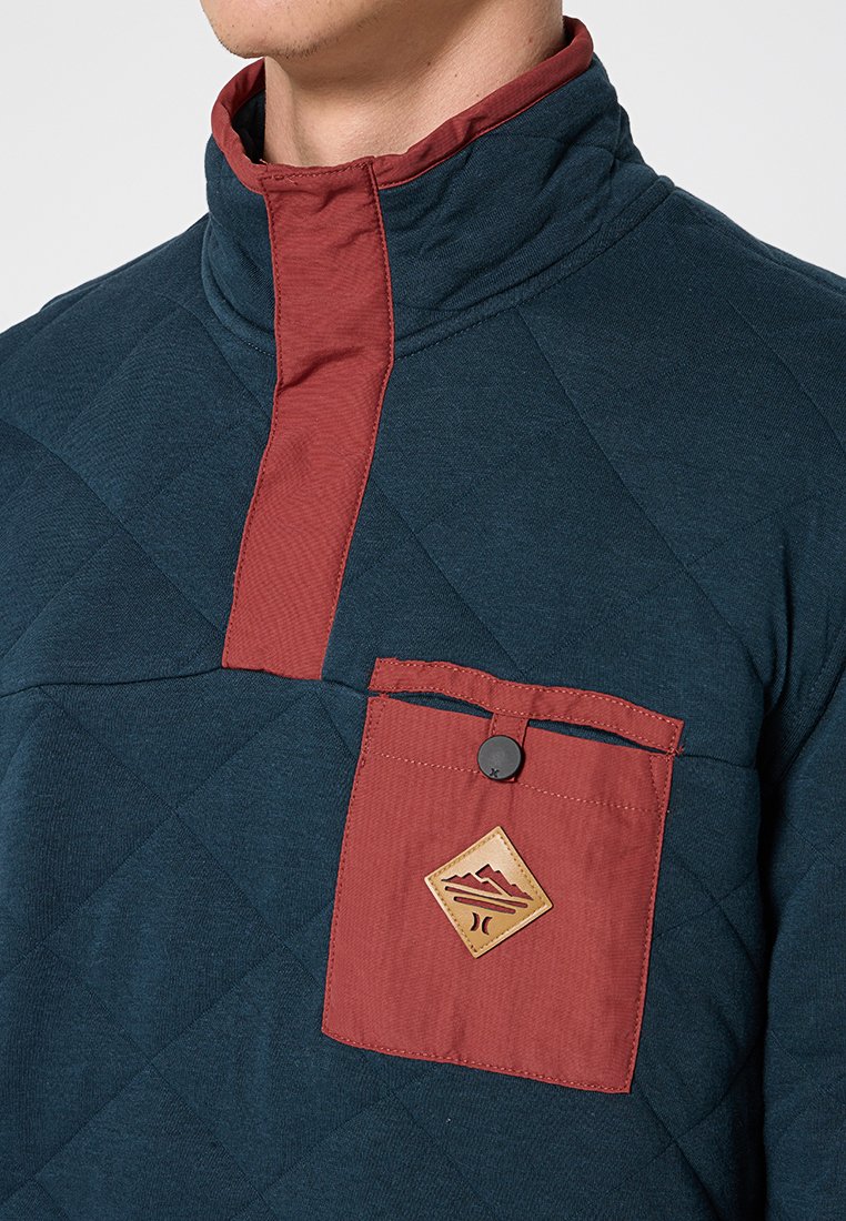 Maglione imbottito in verde scuro con colletto rosso e tasca sul petto, caratterizzato da una chiusura a bottoni e un'etichetta con logo a contrasto sulla tasca.