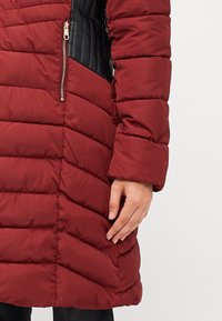 Sarkans puffa mētelis ar horizontālu quilted dizainu, ar melnu sānu paneļu un zelta rāvējslēdzēja kabatu. Gluda tekstūra, pilna piedurkne.