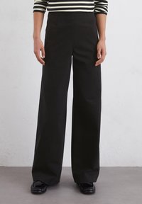 Zwarte wijde broek van gladde stof, met een hoge taille en gestructureerd ontwerp, gecombineerd met zwarte loafers.