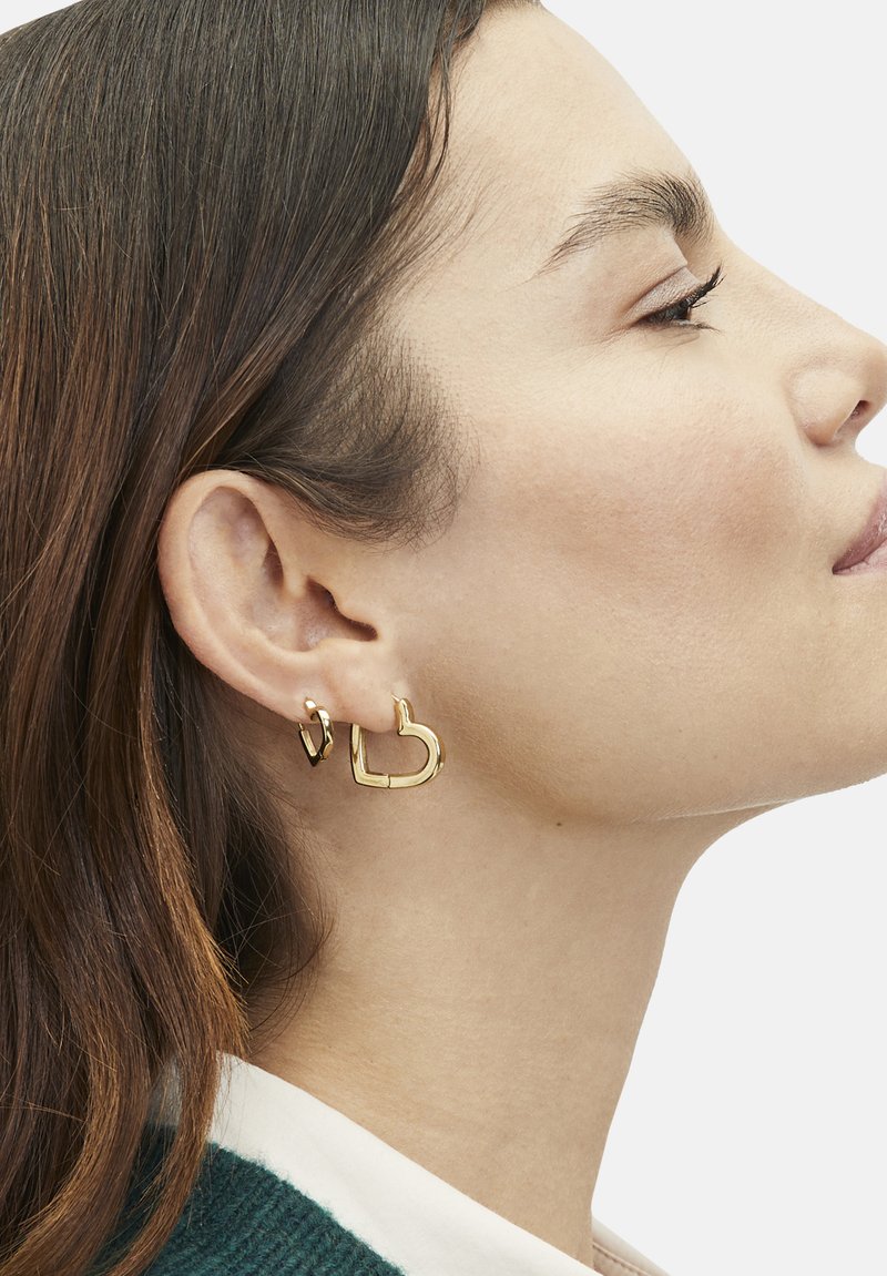 Gold hoop earrings in heart shapes, featuring a shiny finish. The earrings vary in size, with one larger than the other, worn on the left ear.
