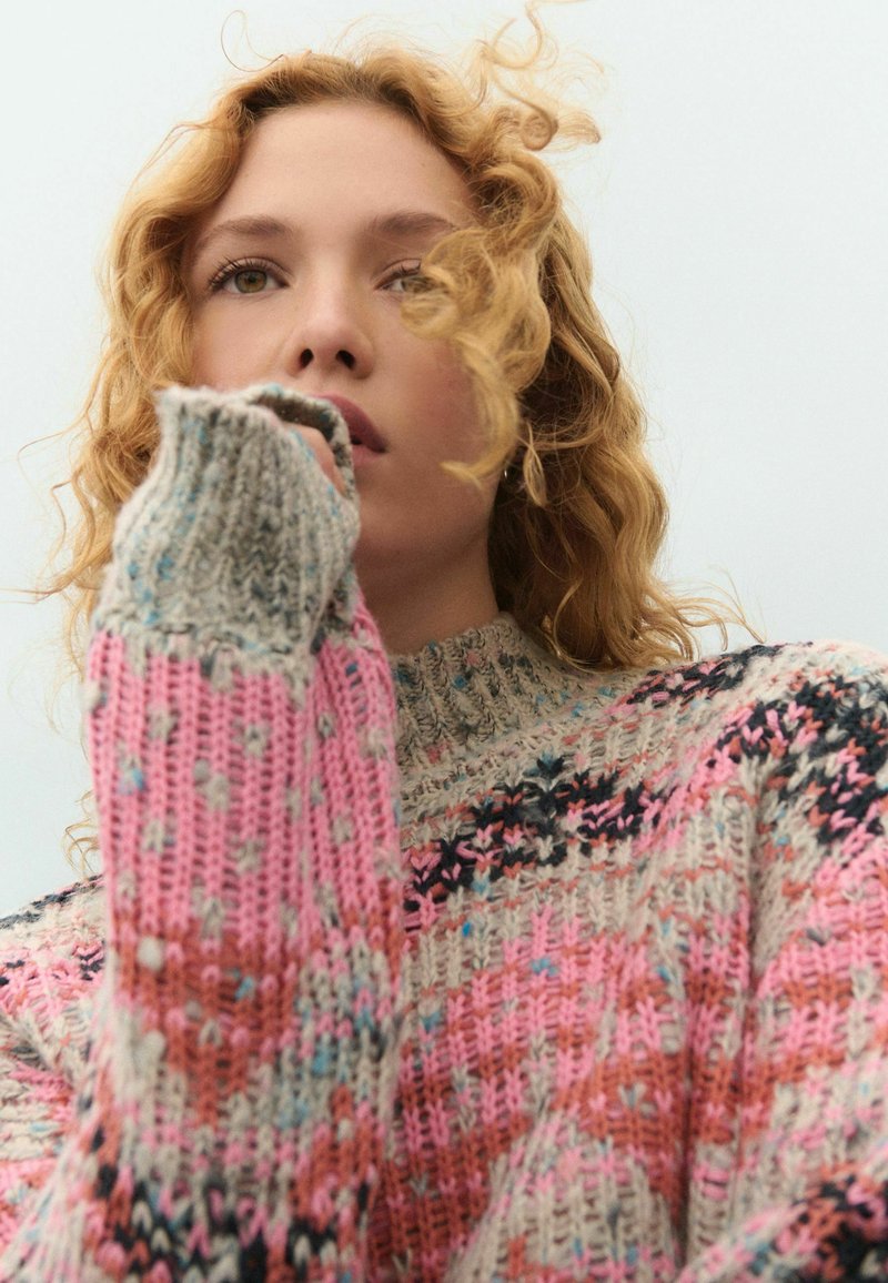 Pull en laine présentant des teintes rose, beige et gris avec un motif texturé. Col montant et manches longues avec des détails de points variés.