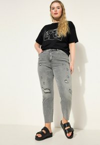 Camiseta negra con diseño gráfico contorneado, combinada con jeans skinny gris desgastados que presentan desgarros y deshilachados. Sandalias negras de tipo slide.