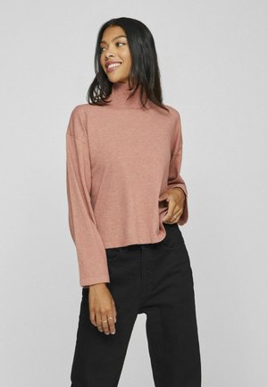 Pull turtleneck rose clair avec des manches longues et amples ; associé à un pantalon noir. Texture douce, coupe décontractée et design simple.