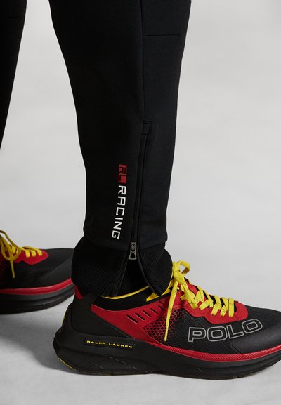 Pantalons de sport noirs avec le logo "RL RACING" rouge et des poignets zippés, accompagnés de baskets noires et rouges avec des lacets et des accents jaunes.