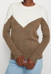 Brązowo-kremowy sweter z wzorem w jodełkę, wykonany z miękkiego dzianinowego materiału, z okrągłym dekoltem i długimi rękawami.