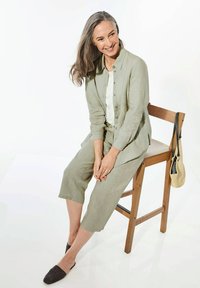 Leicht olivgrünes Leinenoutfit mit einem kurzärmeligen Hemd mit Kragen und verkürzten Hosen. Schwarze gewebte Slipschuhe. Einfacher Holzstuhl, beige Tasche.