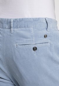 Ljust blå kordflodbyxor med vertikala ribbingar, med en svart knapp och en bakficka med synlig sömdetalj.