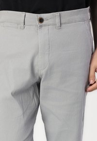 Pantalones de algodón gris claro con una textura suave, que presentan un cierre de botón y bolsillos frontales. La cintura está cosida para mayor durabilidad.