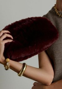 Bourgondische nepbont clutch vastgehouden in de hand, ovale vorm, zachte textuur. Draagt een gebreide grijze top en gouden armbanden, met een chunky ontwerp.