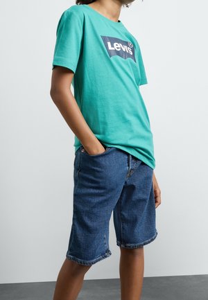 Persona che indossa una maglietta Levi's turchese e pantaloncini di jeans blu fino al ginocchio, con una mano in tasca, in piedi contro uno sfondo semplice.