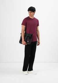 Burgundi színű pólót, fekete nadrágot és fehér sneakers-t viselő férfi fekete Tommy Jeans vászontáskát tart a kezében, egy fehér háttér előtt áll.