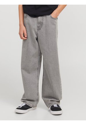 Person iført løstsiddende grå jeans og sorte sneakers med hvide såler, stående med den ene hånd i lommen mod en ensfarvet baggrund.