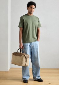 T-shirt verde de algodão, jeans azuis largos de ganga, e uma bolsa de couro bege com detalhes castanhos e duas alças, sobre um fundo claro.