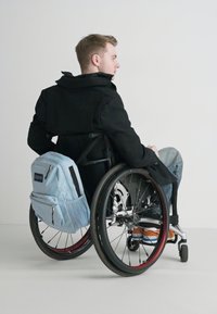 Blue backpack with black accents, zipper pockets, and textured fabric. Black coat, wheelchair with red rims, and visible wheels. Casual jeans and shoes.