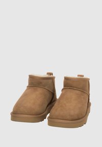 Para brązowych zamszowych botków za kostkę z miękkim białym wykończeniem i gumowymi podeszwami, pokazanych na jednolitym jasnoszarym tle.