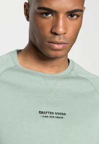 Lys grøn sweatshirt med rund hals, prydet med et sort tekstlogo, der står "Crafted Goods - Pier One Denim." Glat, blød tekstur.