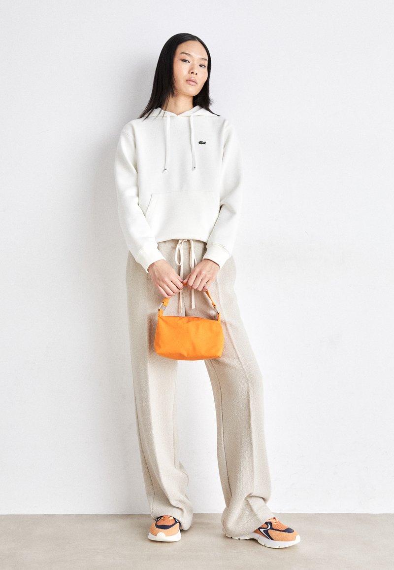 Biała bluza z kapturem ze sznurkiem, jasnobeżowe szerokie spodnie, pomarańczowa torebka oraz sneakersy z pomarańczowymi i granatowymi akcentami. Widoczne teksturowane tkaniny.