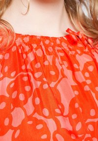 Blouse orange vif en tissu léger et doux avec des motifs circulaires roses. L'encolure est élastiquée avec des liens à nouer.