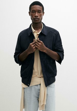 Mann trägt eine marineblaue Jacke über einem beigen Poloshirt mit einem beigen Pullover, der um die Taille gebunden ist, und hellblaue Jeans, steht vor weißem Hintergrund.