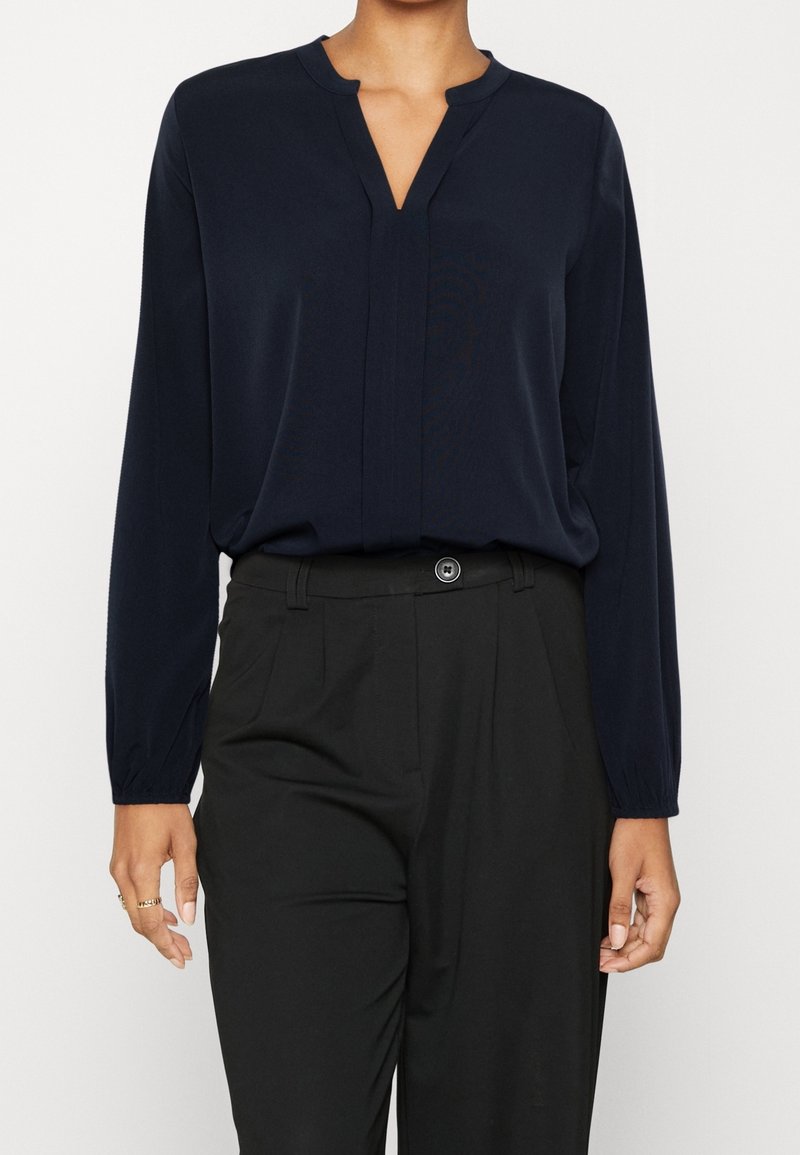 Femme portant un chemisier bleu marine à manches longues avec un col en encoche en V, rentré dans un pantalon noir taille haute et ajusté.