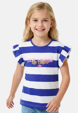 Fille blonde souriante portant une chemise à rayures bleues et blanches avec des manches à volants et le texte "East London Lee Cooper", debout devant un fond blanc.