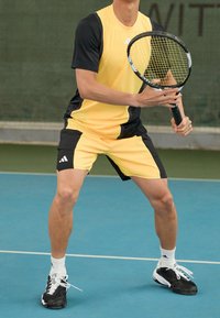 Conjunto deportivo amarillo y negro con mangas cortas y pantalones cortos. El jugador sostiene una raqueta de tenis en una cancha azul, vistiendo zapatillas de tenis negras.