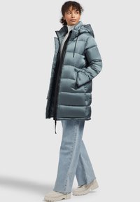 Palton captusit dintr-un material moale de culoare teal cu glugă, fermoar frontal, două buzunare laterale și accente negre. Combinat cu blugi ușori din denim evazați.