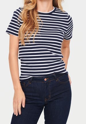 Mujer con camiseta de manga corta a rayas azul marino y blanca, metida dentro de unos vaqueros de mezclilla azul oscuro, con una mano en el bolsillo y la otra relajada al costado.