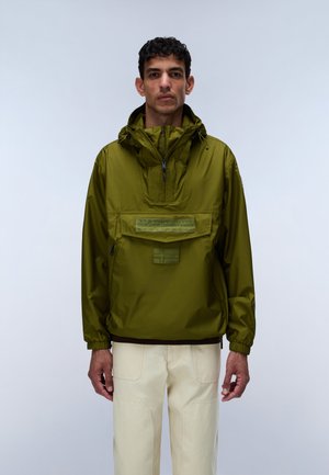 Mann trägt eine olivgrüne Kapuzenjacke mit Fronttasche und cremefarbene Hose, steht vor einem einfarbigen hellen Hintergrund.