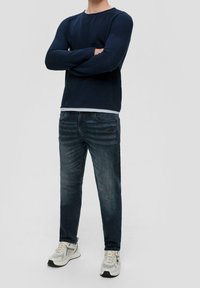 Mies, który ma na sobie granatowy, teksturowany sweter z długim rękawem, ciemne, lekko przetarte dżinsy i białe sneakersy, stoi z założonymi rękami.