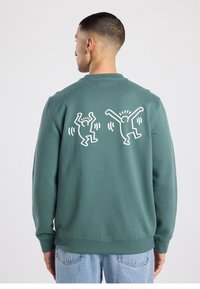 Green sweatshirt made of soft fabric with a crew neckline and ribbed cuffs. Features two white graphic figures on the back, mid-action.