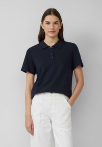 Granatowa koszulka polo z krótkim rękawem i kołnierzykiem, z zapięciem na guziki. Noszona z białymi spodniami w szerokim kroju, o gładkiej fakturze tkaniny.
