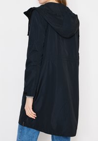 Femme portant un long imperméable noir à capuche avec une taille élastique et une poche sur la manche, associée à un jean bleu, vue de dos.