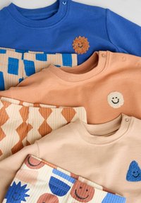 Színes gyermekruhákat tartalmazó kollekció, kék felső oroszlán hímzéssel, barack színű felsők, és geometrikus mintákkal ellátott mintás nadrágok.