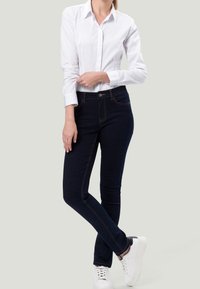 Weißes Hemd mit Knöpfen, in dunkle blaue Skinny-Jeans gesteckt, mit orangefarbenen Nähdetails, kombiniert mit weißen Sneakers auf hellem Hintergrund.
