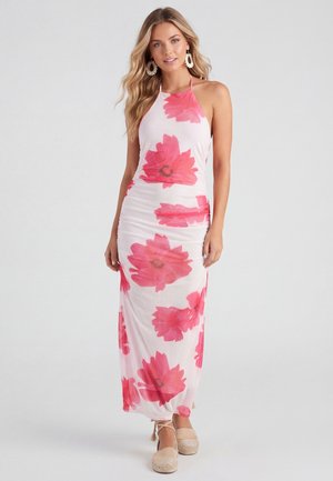 Kvinde iført en hvid halterneck maxikjole med store pink blomsterprint og beige espadrillesko, poserer mod en ensfarvet baggrund.