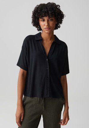 Donna con capelli scuri e ricci che indossa una camicia nera larga a maniche corte con bottoni e pantaloni verde oliva texture su uno sfondo semplice.