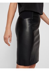 Jupe en cuir noir à texture lisse, avec une couture latérale, une coupe ajustée et une ceinture élastique pour le confort.