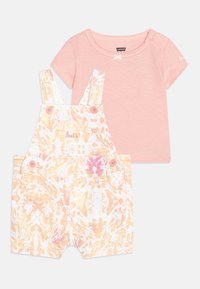 Jasnoróżowa koszulka z krótkim rękawem w zestawie z białymi ogrodniczkami w wzór tie-dye w odcieniach żółtego i różowego, z różowymi guzikami oraz kieszeniami.