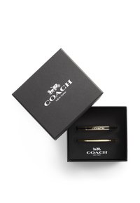 Svart presentask med texturerad yta innehåller två slipsklämmor i guldton. Asken har "COACH NEW YORK"-logotyp i vitt på toppen och inuti.