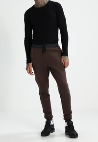 Camisa negra de manga larga y pantalones tipo jogger marrones con cintura elástica. Los pantalones presentan un diseño de pierna cónica y se combinan con zapatillas negras.