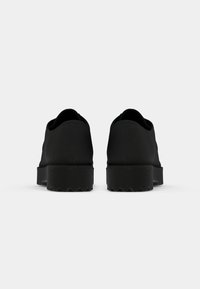 Chaussures noires en caoutchouc avec une surface lisse, un bout rond et une semelle épaisse et texturée. Conçues sans lacets, elles présentent une silhouette minimaliste.