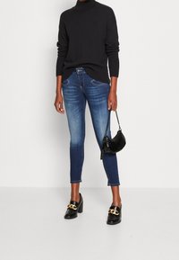 Femme portant un col roulé foncé, un jean skinny bleu délavé, des mocassins à talons noirs ornés d'une chaîne dorée, tenant un petit sac à main noir avec une bandoulière chaîne.