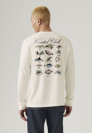 Oseba s kratkimi blond lasmi, oblečena v belo dolgo majico z barvitimi obalnimi morskimi živalmi in napisom "Coastal Club" na hrbtni strani.