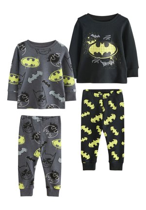 To unge drenge med blondt hår sidder på en seng iført matchende Batman-inspirerede pyjamas, smilende og kigger lidt mod højre.