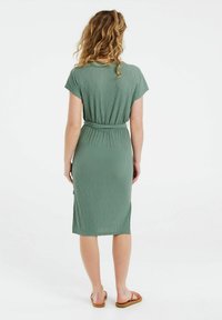 Vestido verde acanalado con mangas cortas, cintura ceñida con cinturón y diseño de longitud media. Presenta una textura simple y suave. Combinado con sandalias planas.