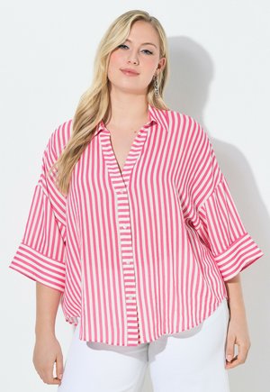 Frau trägt ein locker sitzendes pink-weiß vertikal gestreiftes Hemd mit weiten Ärmeln und weiße Hose, steht vor weißem Hintergrund.