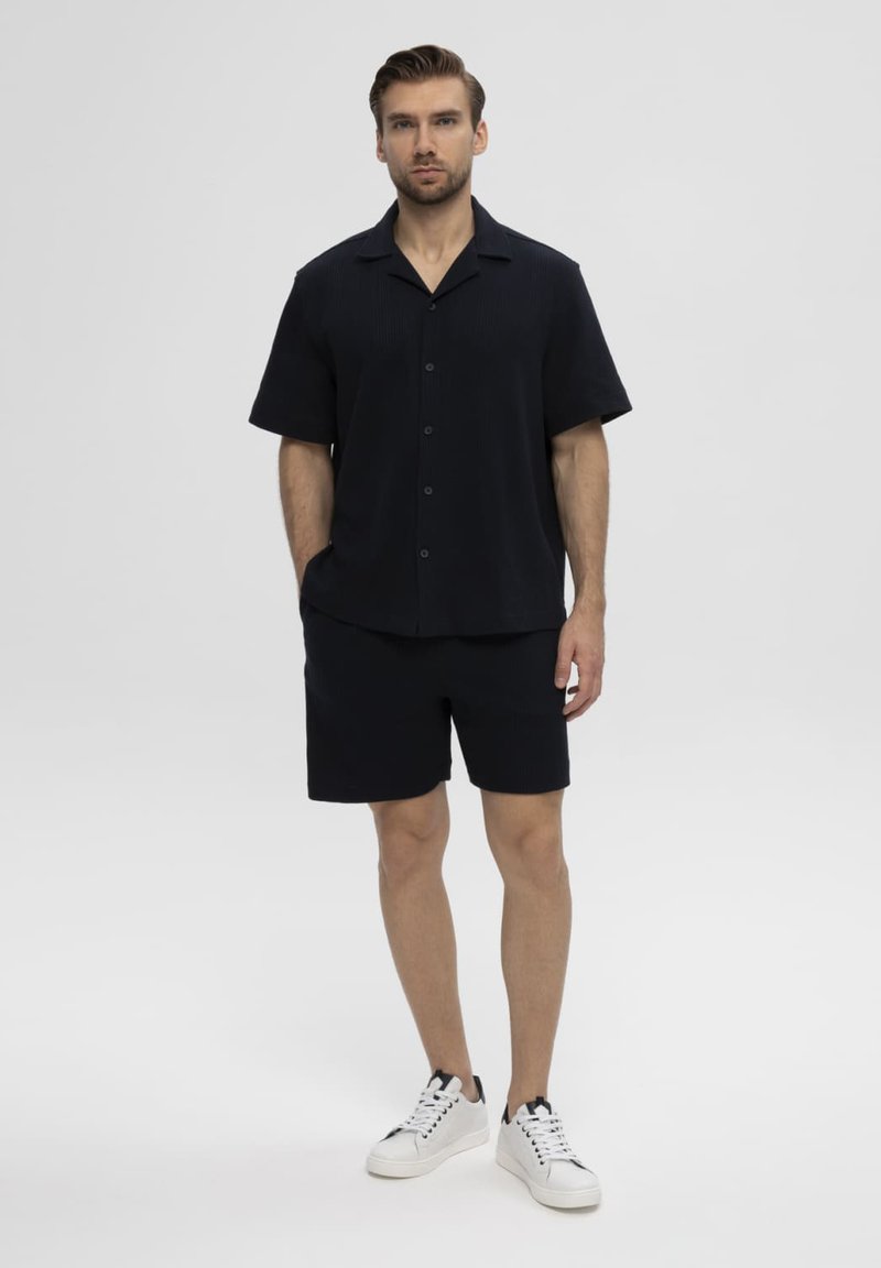 Homme portant une chemise noire à manches courtes boutonnée et un short noir, debout devant un fond clair uni, les mains dans les poches.